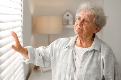 Photo of Loneliness concept. Sad senior woman looking through window blinds at home