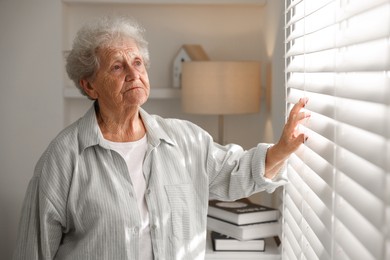 Photo of Loneliness concept. Sad senior woman looking through window blinds at home