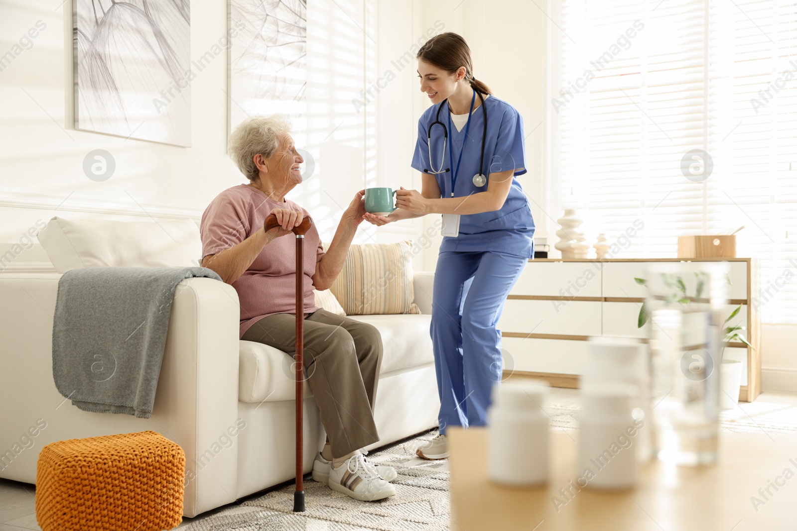 Photo of Caregiver giving cup of drink to senior woman indoors. Home health care service