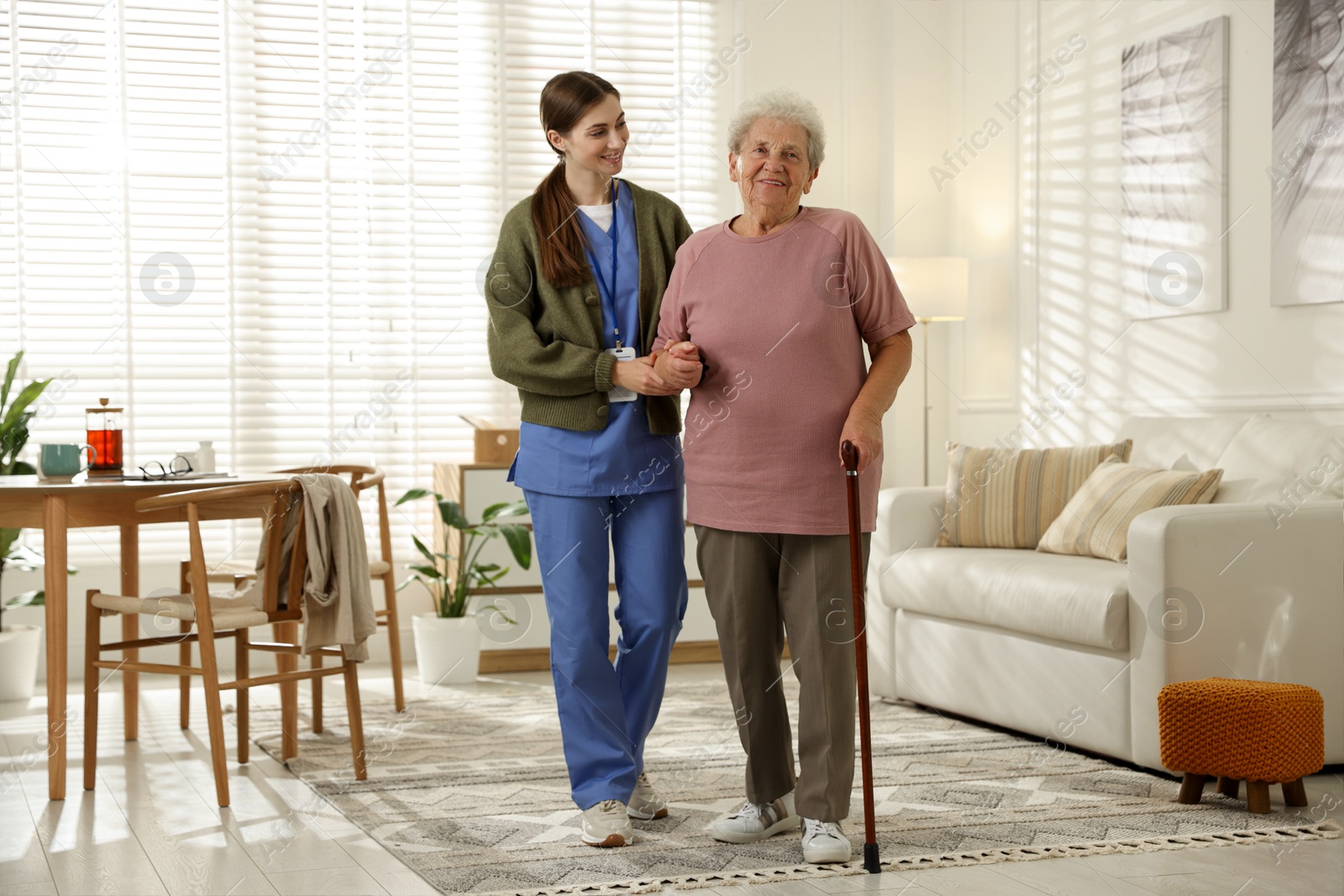 Photo of Caregiver assisting senior woman with walking cane indoors. Home health care service