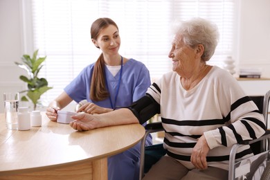 Photo of Caregiver measuring patient's blood pressure at table indoors. Home health care service