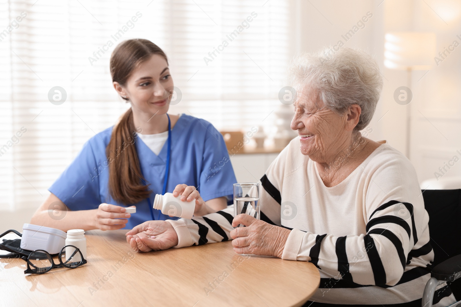 Photo of Caregiver giving pills to senior woman at table indoors. Home health care service