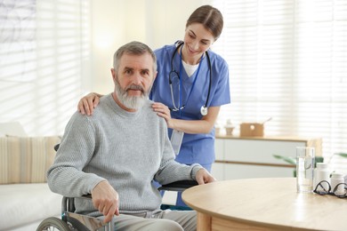 Photo of Caregiver assisting senior man in wheelchair indoors. Home health care service