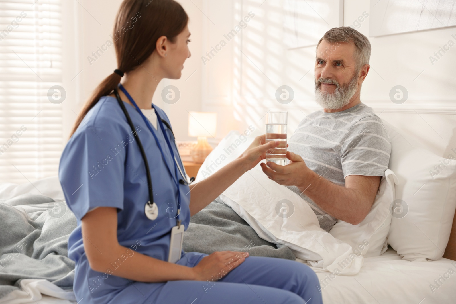 Photo of Caregiver giving water to senior man on bed indoors. Home health care service