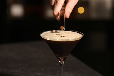 Photo of Bartender putting coffee bean into glass with delicious espresso martini at dark table, closeup