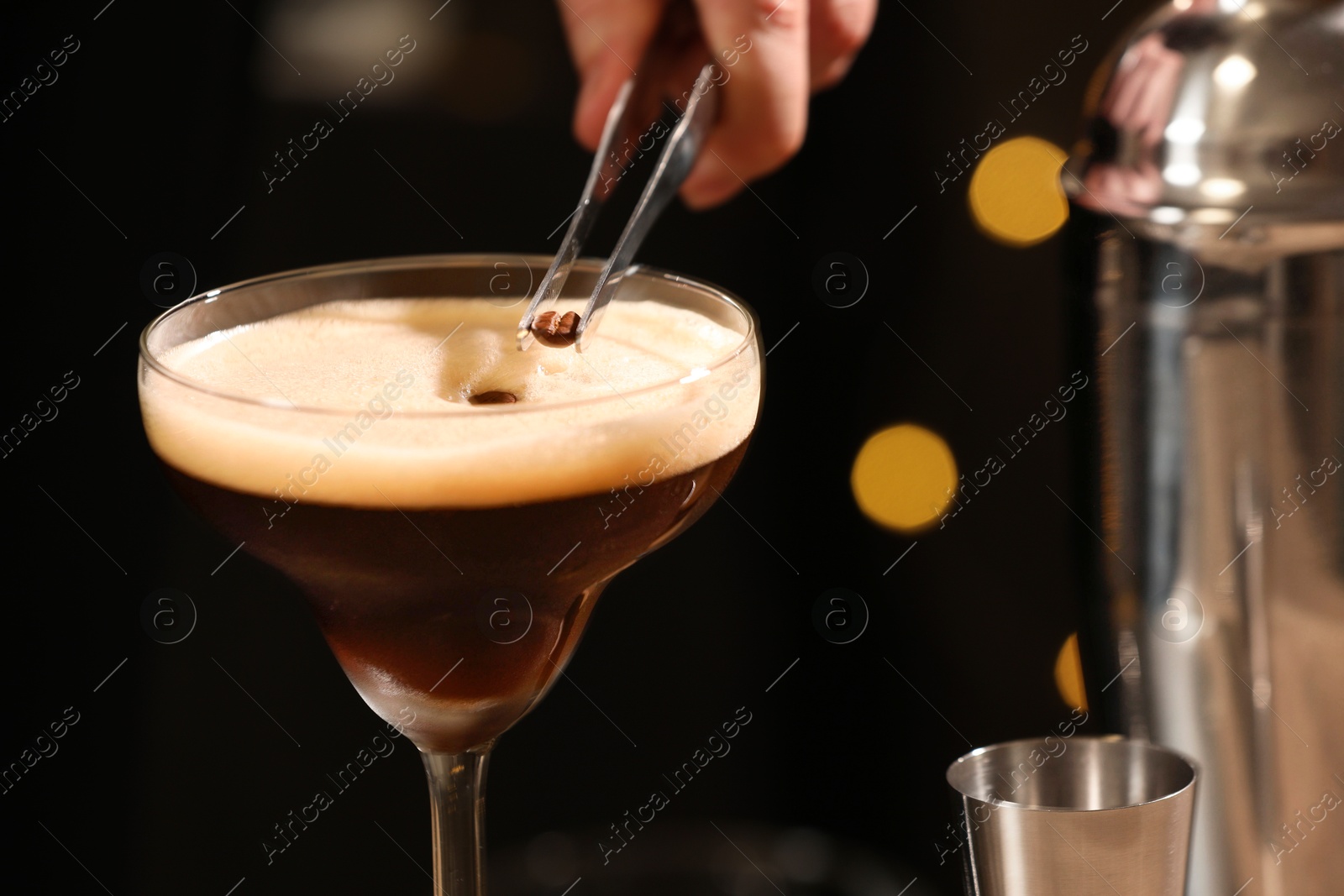 Photo of Bartender putting coffee bean into glass with delicious espresso martini against blurred lights, closeup