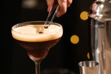 Photo of Bartender putting coffee bean into glass with delicious espresso martini against blurred lights, closeup