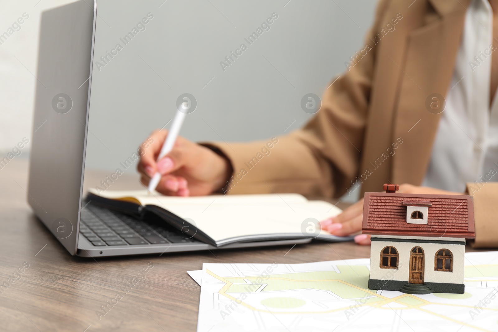 Photo of House hunting. Woman with laptop, papers, notebook and house figure at wooden table, closeup