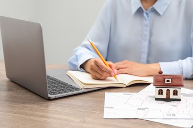 Photo of House hunting. Woman with laptop, papers, notebook and house figure at wooden table, closeup