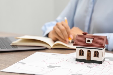 Photo of House hunting. Woman with laptop, papers, notebook and house figure at wooden table, closeup