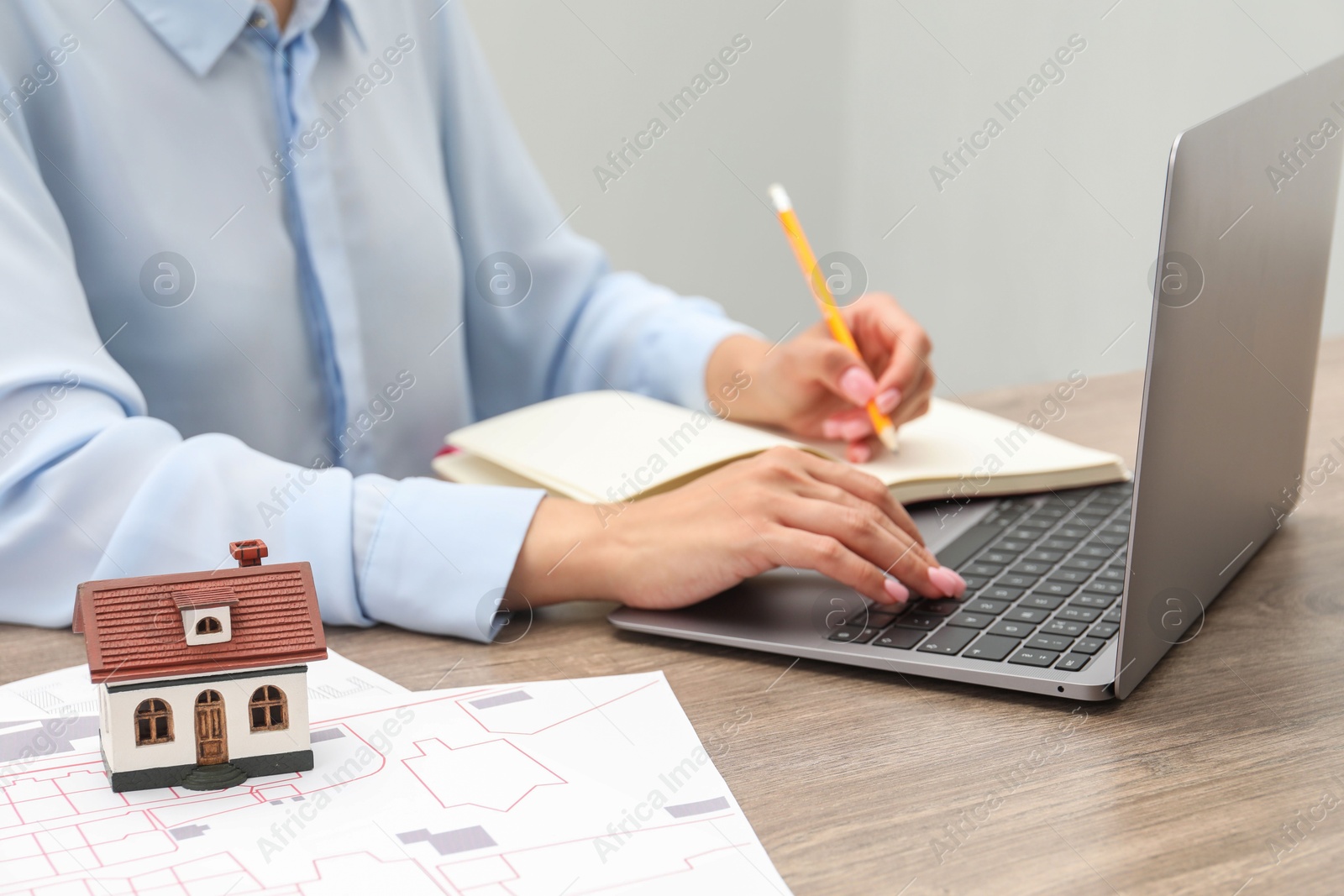 Photo of House hunting. Woman with laptop, papers, notebook and house figure at wooden table, closeup