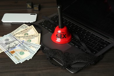 Photo of Sex work. Laptop, mask, money, smartphone, dice and bell on wooden surface, closeup