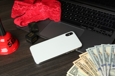 Photo of Sex work. Laptop with lingerie, smartphone, bell, dice and money on wooden surface, closeup