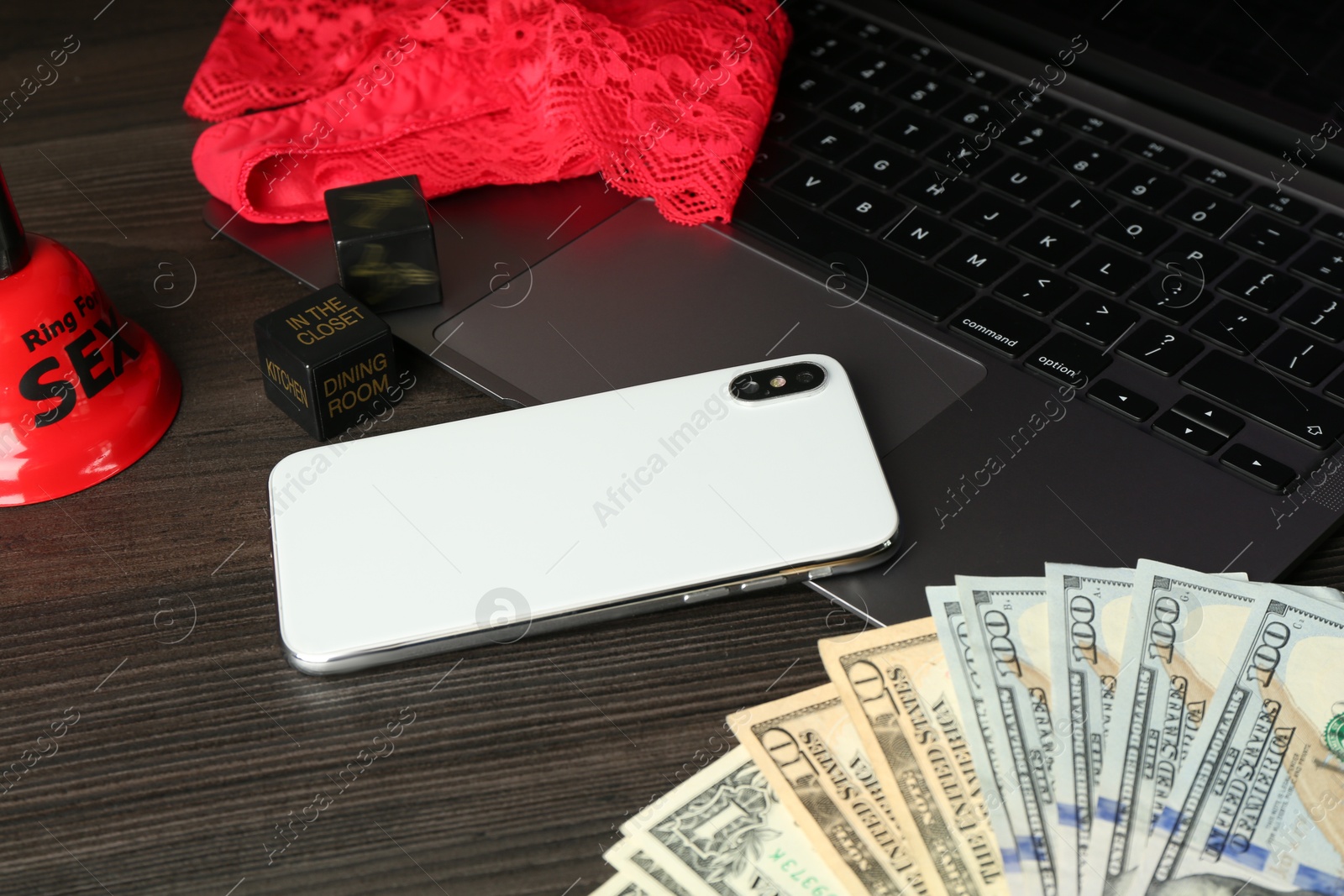 Photo of Sex work. Laptop with lingerie, smartphone, bell, dice and money on wooden surface, closeup