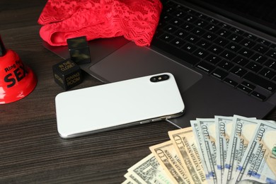 Sex work. Laptop with lingerie, smartphone, bell, dice and money on wooden surface, closeup