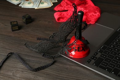 Photo of Sex work. Laptop, lingerie, bell, mask and money on wooden surface, closeup