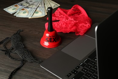 Photo of Sex work. Laptop, lingerie, bell, mask and money on wooden surface, closeup