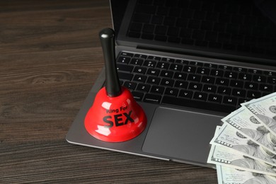 Photo of Sex work. Laptop with bell and money on wooden surface, closeup