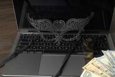 Photo of Sex work. Laptop with mask and money on wooden surface, closeup