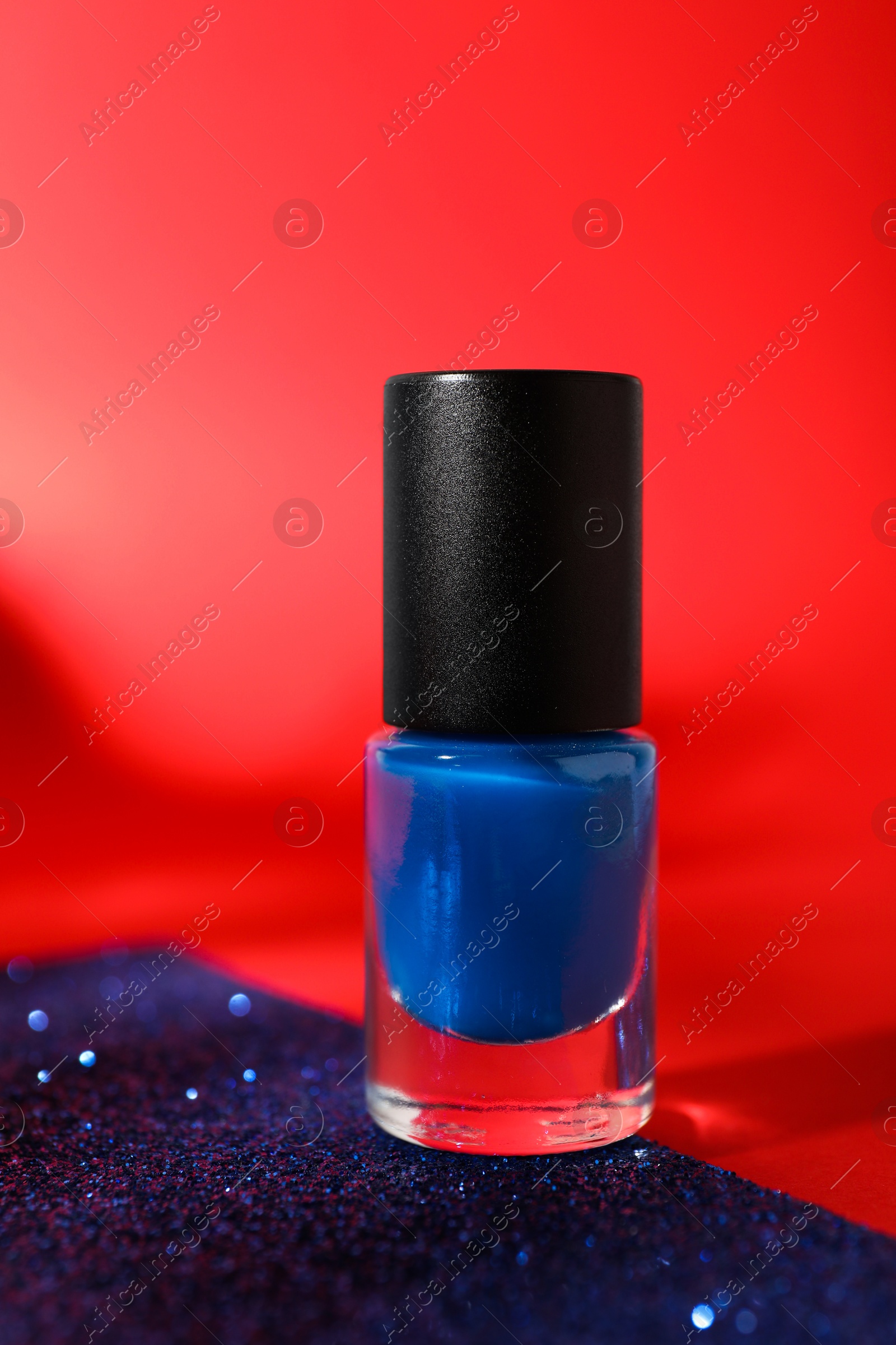 Photo of Blue nail polish in bottle on bright glitter surface against red background