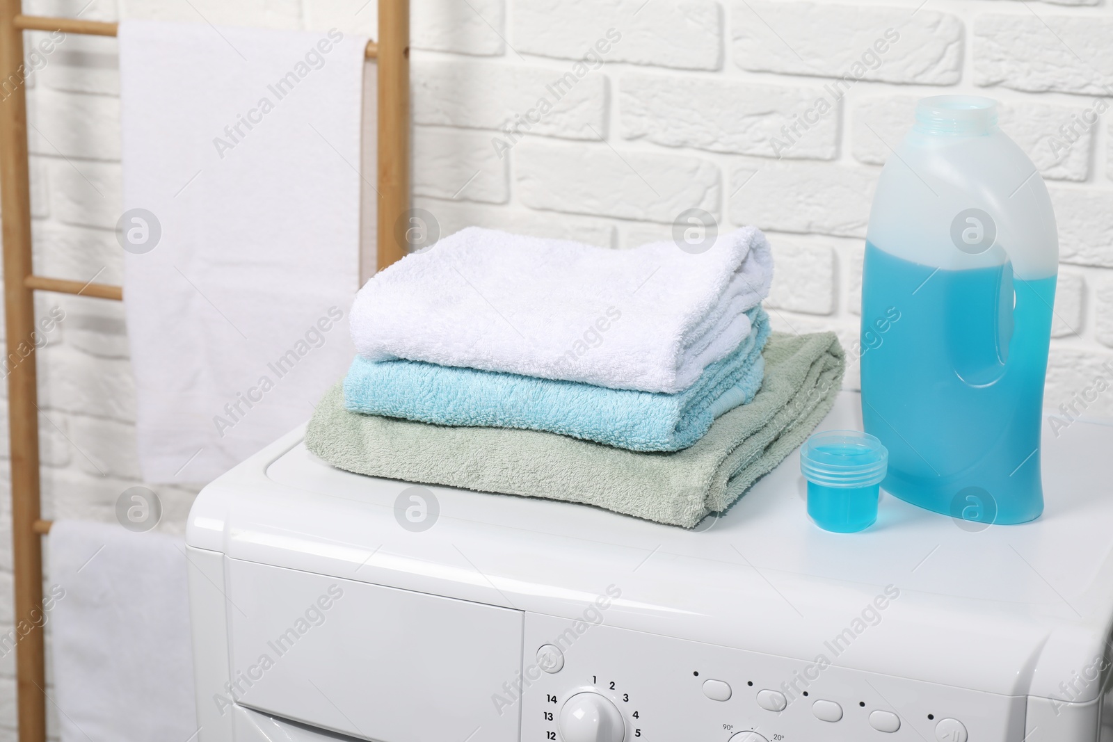 Photo of Laundry detergent, cap and stacked clean towels on washing machine in bathroom