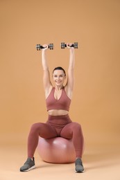 Photo of Woman exercising with dumbbells on fitball against beige background