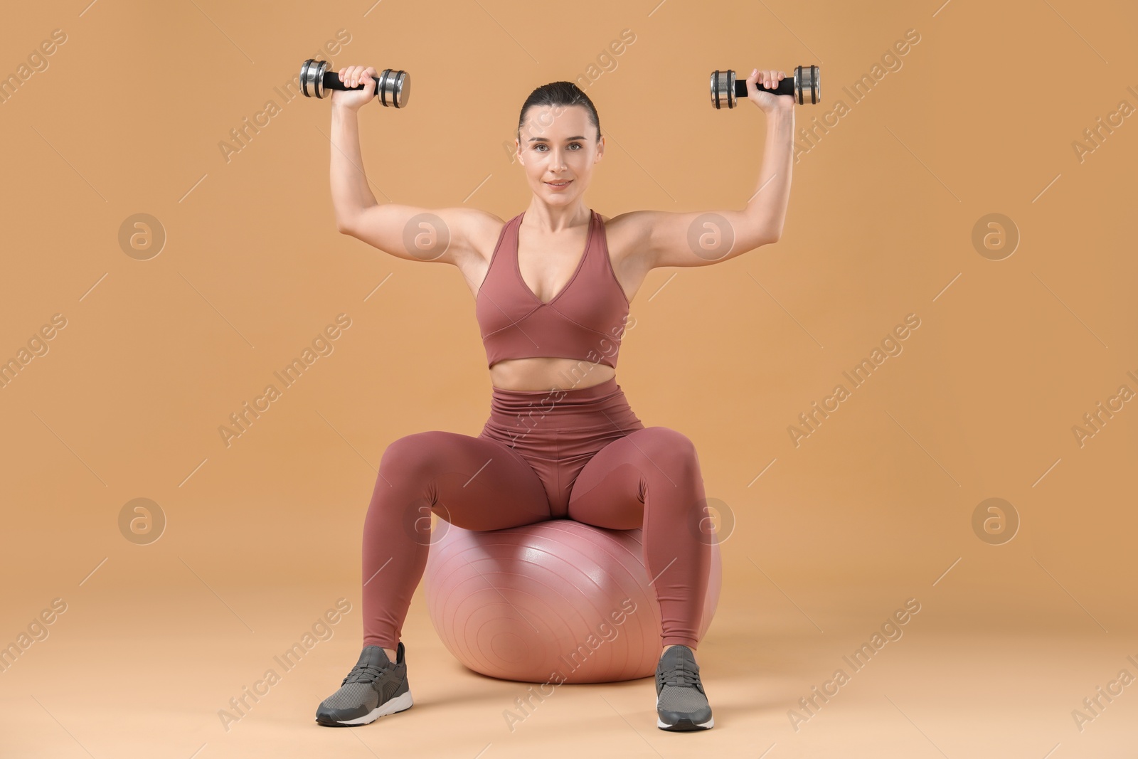 Photo of Woman exercising with dumbbells on fitball against beige background