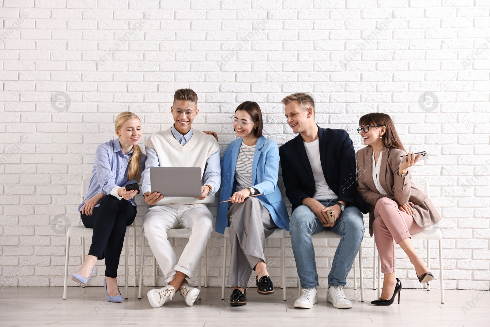 Photo of Group of people using different gadgets on chairs near white brick wall indoors. Modern technology