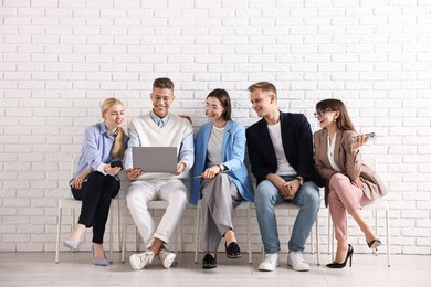 Photo of Group of people using different gadgets on chairs near white brick wall indoors. Modern technology