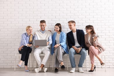 Photo of Group of people using different gadgets on chairs near white brick wall indoors. Modern technology