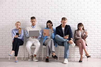 Group of people using different gadgets on chairs near white brick wall indoors. Modern technology