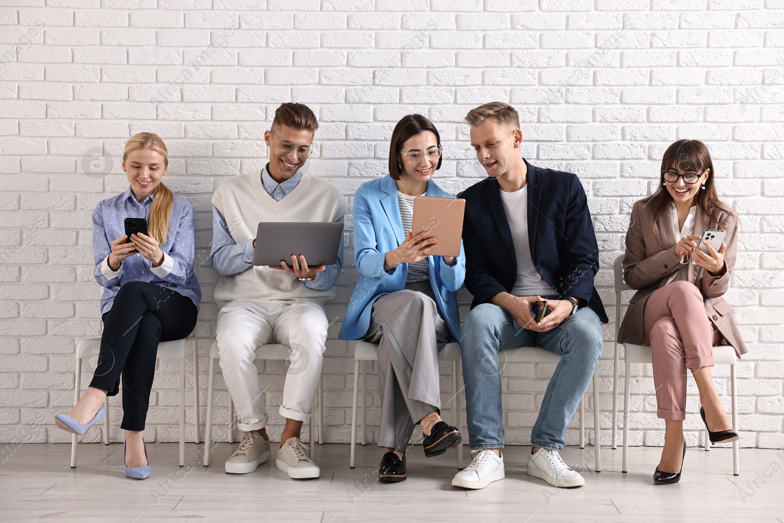 Photo of Group of people using different gadgets on chairs near white brick wall indoors. Modern technology