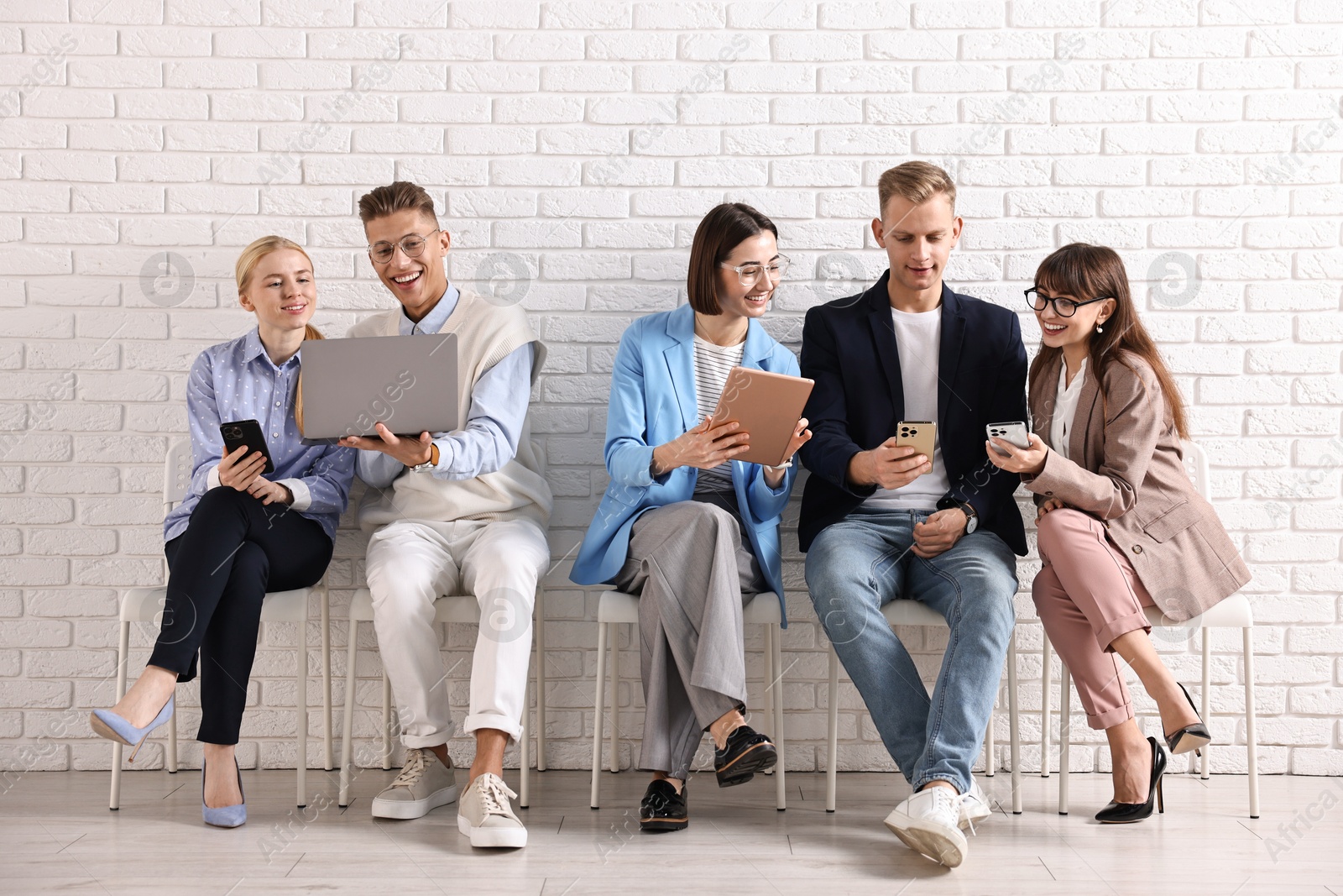 Photo of Group of people using different gadgets on chairs near white brick wall indoors. Modern technology