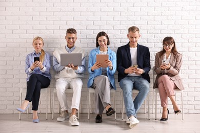 Photo of Group of people using different gadgets on chairs near white brick wall indoors. Modern technology