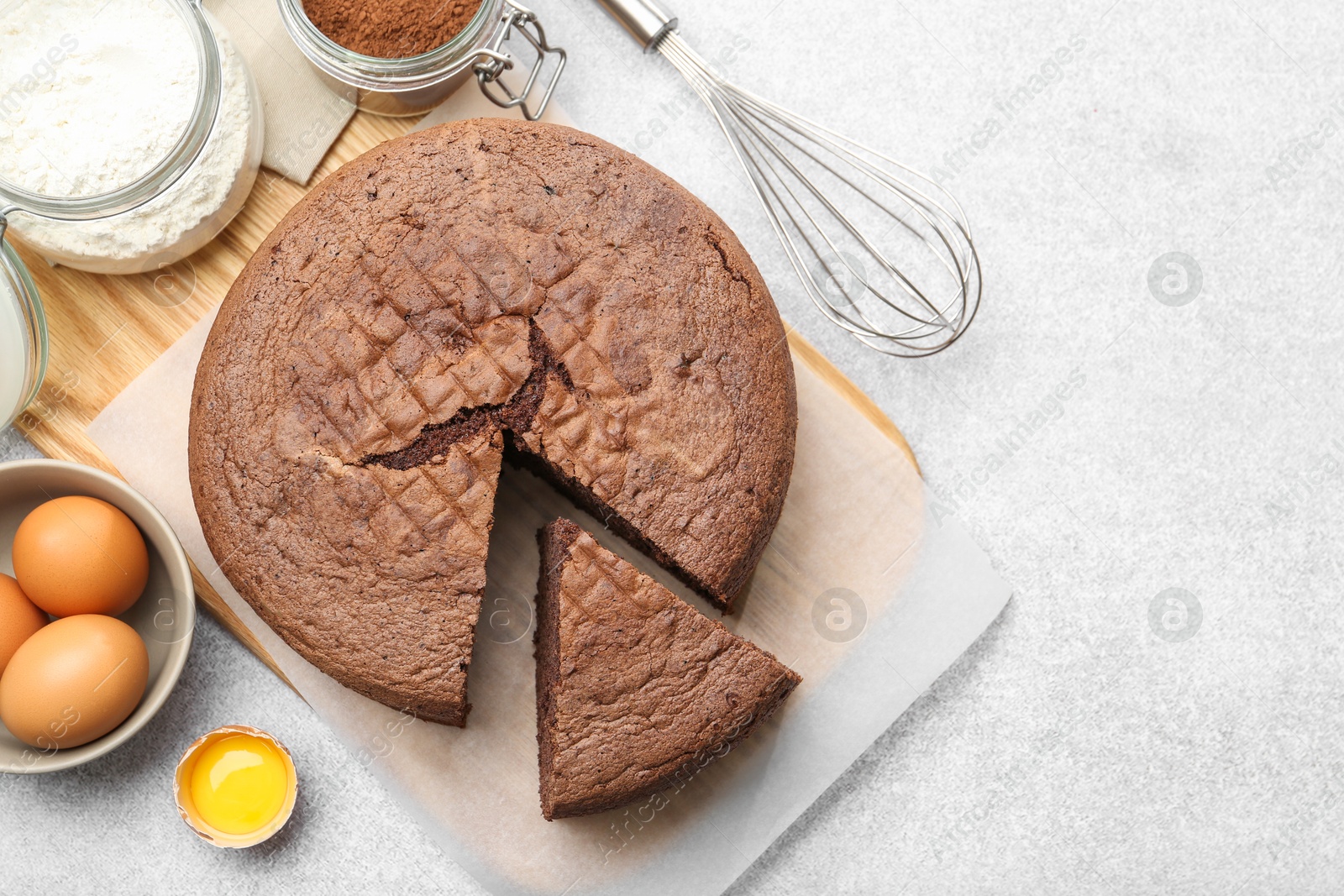 Photo of Cut chocolate sponge cake, whisk and ingredients on light table, flat lay