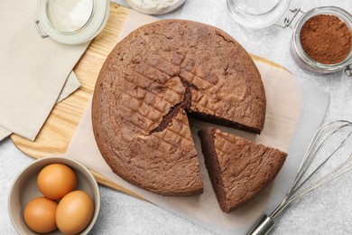 Photo of Cut chocolate sponge cake, whisk and ingredients on light table, flat lay