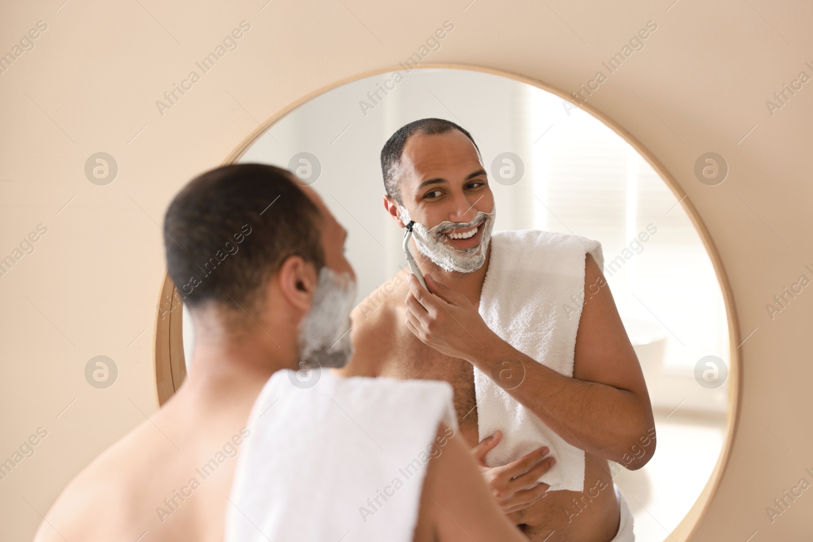 Photo of Smiling man shaving with razor near mirror in bathroom