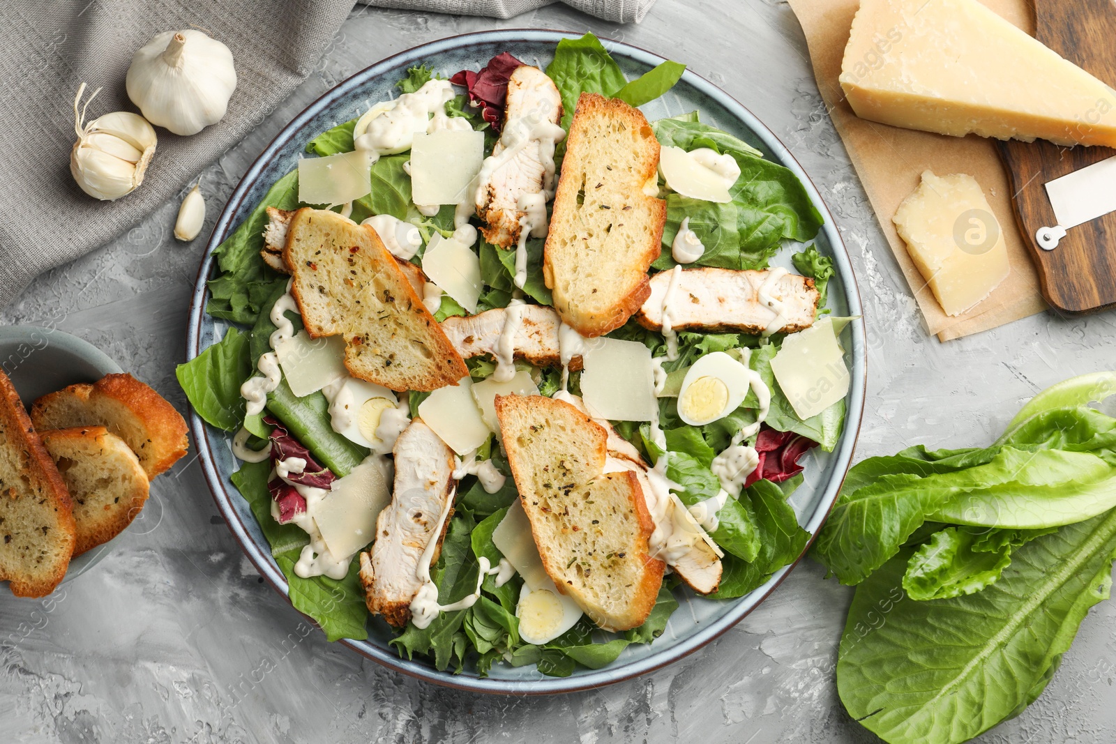 Photo of Tasty Caesar salad with chicken and ingredients on gray textured table, flat lay