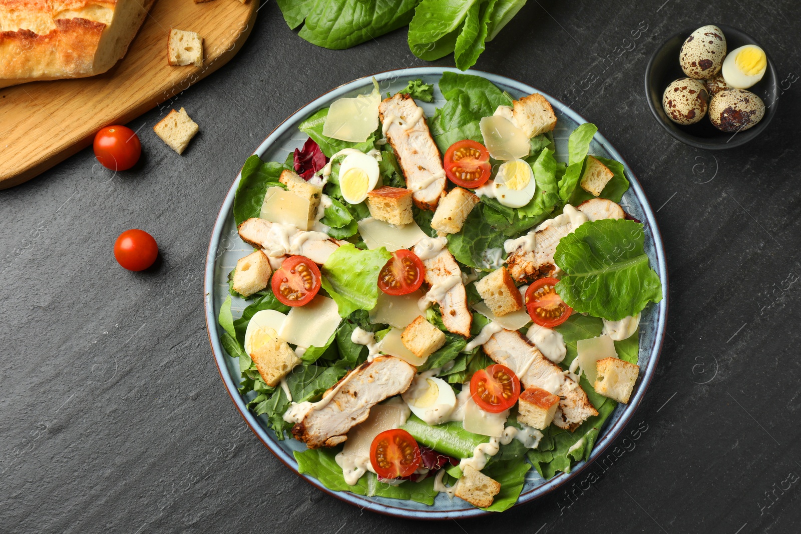 Photo of Tasty Caesar salad with chicken and ingredients on dark gray textured table, flat lay