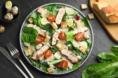 Photo of Tasty Caesar salad with chicken and cutlery on dark gray textured table, flat lay