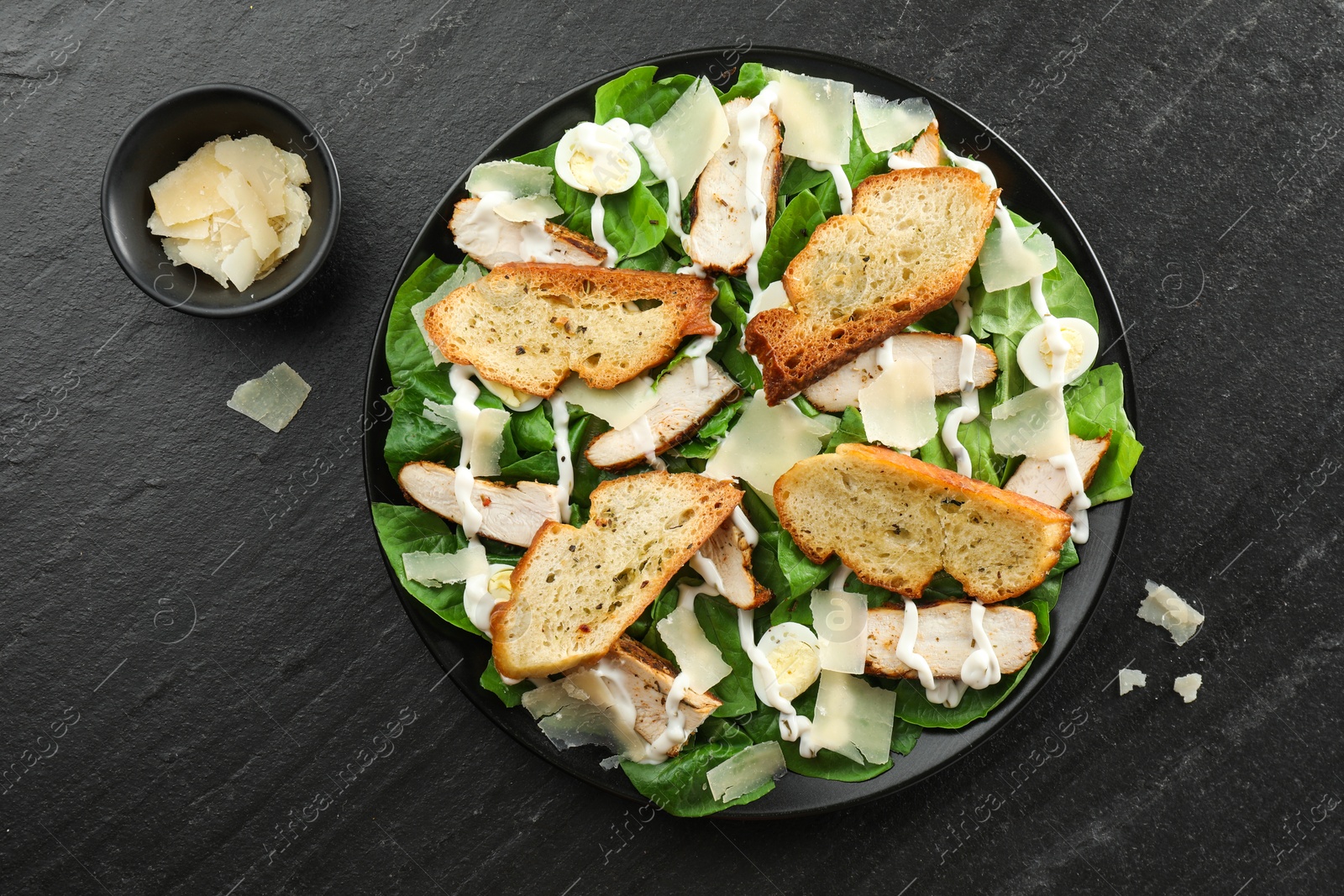 Photo of Delicious Caesar salad with chicken and cheese on black textured table, flat lay