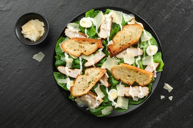 Photo of Delicious Caesar salad with chicken and cheese on black textured table, flat lay