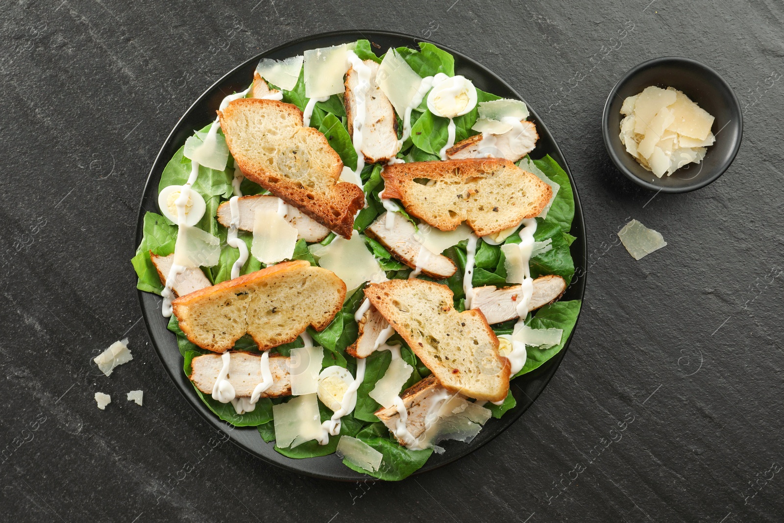 Photo of Delicious Caesar salad with chicken and cheese on black textured table, flat lay