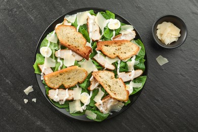 Photo of Delicious Caesar salad with chicken and cheese on black textured table, flat lay