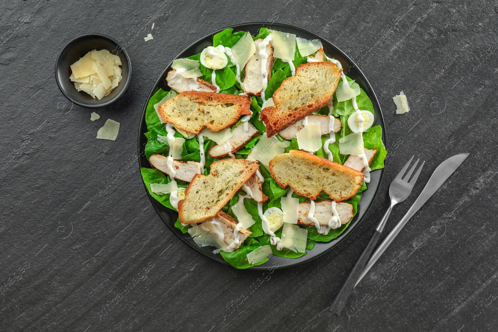 Photo of Delicious Caesar salad with chicken and cheese on black textured table, flat lay