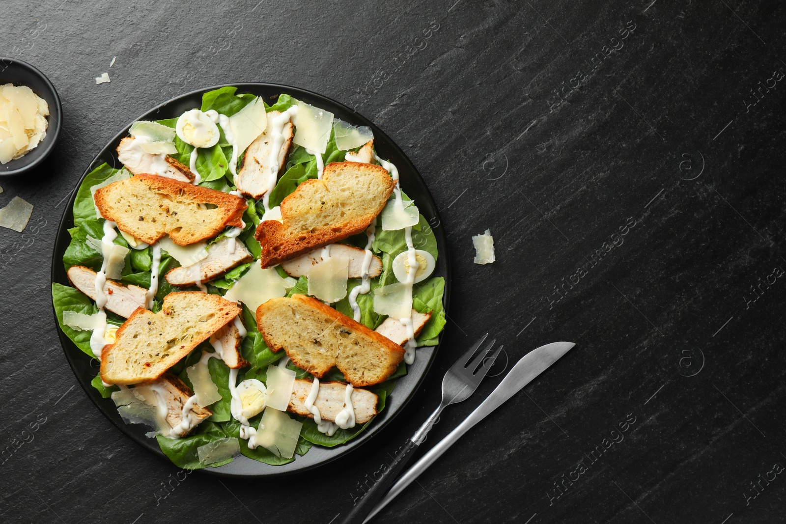 Photo of Delicious Caesar salad with chicken and cheese on black textured table, flat lay. Space for text