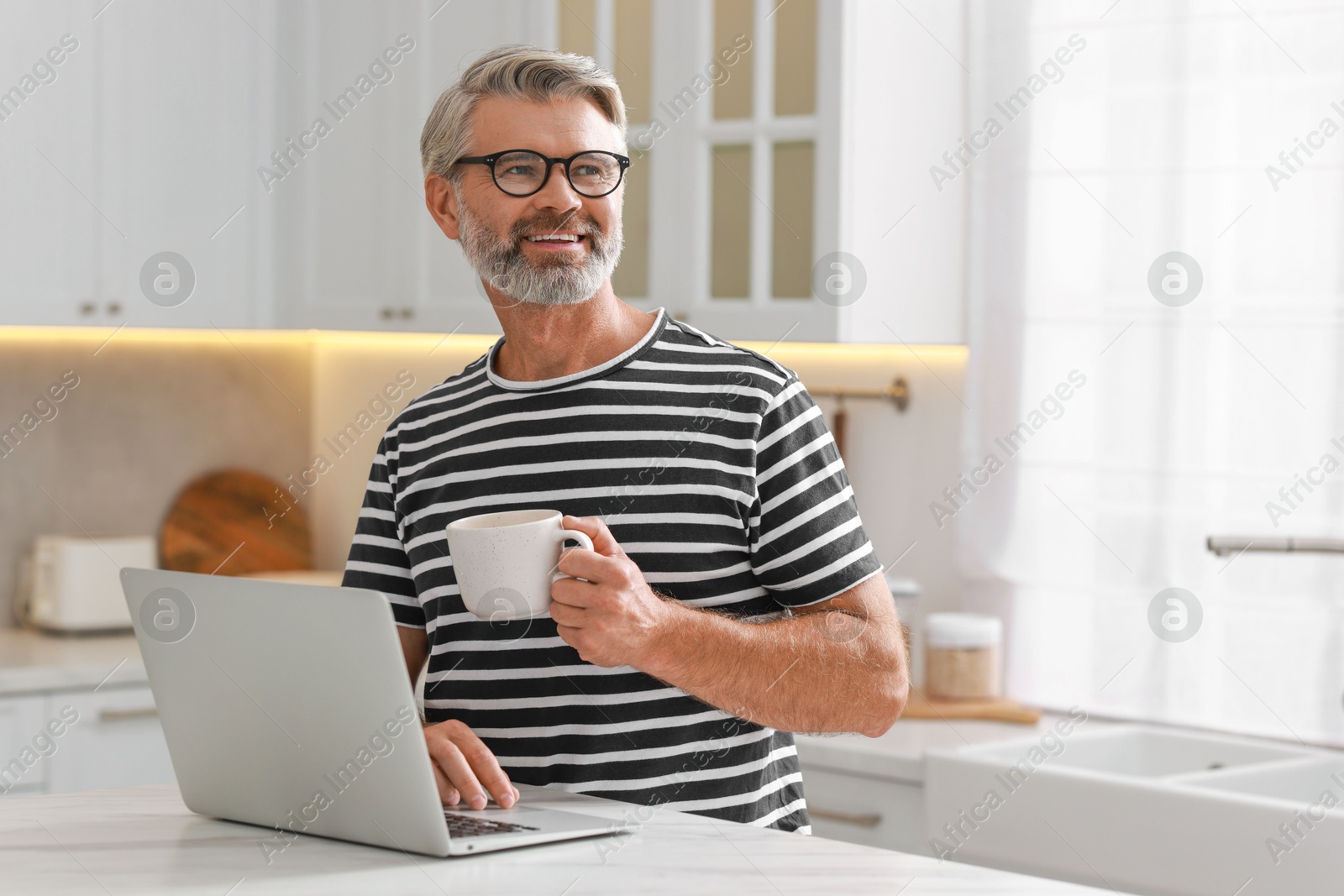 Photo of Happy middle aged man with cup of drink using laptop at white marble table in kitchen