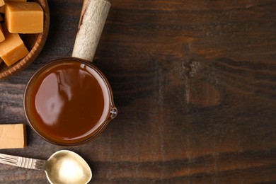 Photo of Tasty caramel sauce in glass measuring cup, sweet candies and spoon on wooden table, flat lay. Space or text