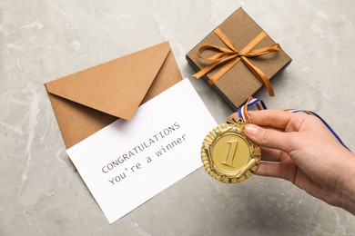 Photo of Woman holding golden medal, card with phrase Congratulations You`re Winner, gift box and envelope at grey marble table, top view