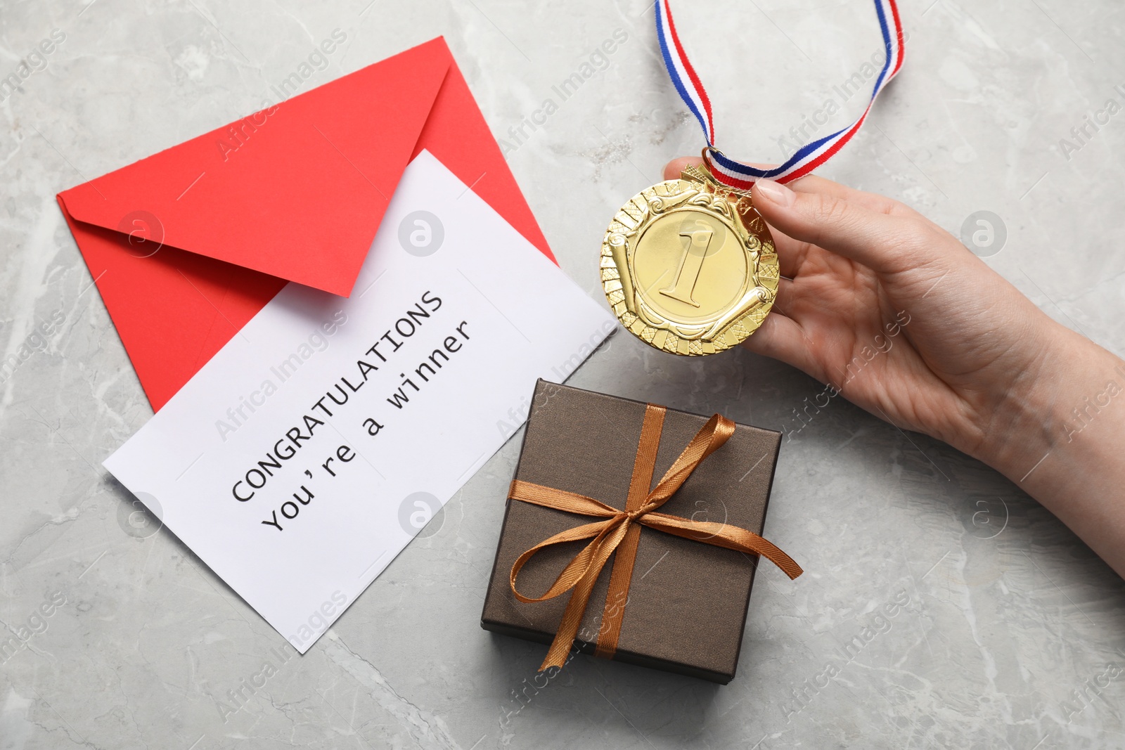 Photo of Woman holding golden medal, card with phrase Congratulations You`re Winner, gift box and envelope at grey marble table, top view